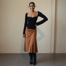 Woman wearing a black long-sleeve top and brown skirt standing against a plain wall.