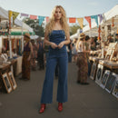 Woman in blue strapless top and jeans standing at an outdoor market with stalls and people in the background.
