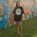 Person wearing a black t-shirt with a white flower design in front of a colorful mural.