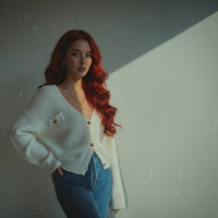 Woman with red hair wearing a white cardigan and blue jeans standing against a dark background.