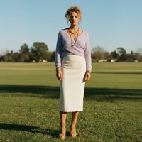 Woman standing on a grassy field wearing a light purple sweater and white skirt.