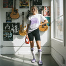 Person wearing a white t-shirt with a graphic design, black shorts, and purple shoes in a room with guitars and posters on the wall.
