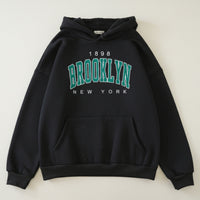 Black hoodie with '1898 Brooklyn New York' text on a gray surface with shoes and a magazine.