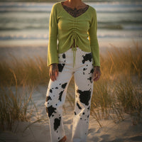 Woman in green sweater and white pants with black spots standing on a sandy beach.