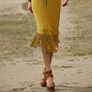 Person wearing a yellow fringe skirt and brown platform shoes on a sandy ground.