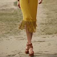 Person wearing a yellow fringe skirt and brown platform shoes on a sandy ground.