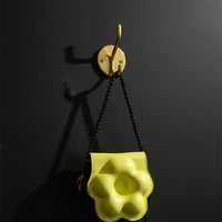 Yellow flower-shaped handbag held by a hand with a black chair and beige cushion in the background.