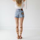 Woman wearing denim shorts with a Sun design in a minimalistic room, wearing wrap sandals and a white tank top