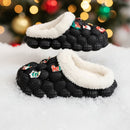 Black slippers with white fuzzy bands on a snowy surface with a Christmas tree in the background