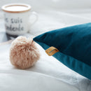 Teal pillow with a beige pom-pom on a white surface, with a blurred coffee mug in the background.