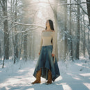 Woman in a long blue skirt and beige top standing in a snowy forest