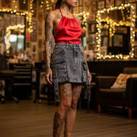 Woman in a red top and denim skirt standing in a tattoo shop with string lights and framed mirrors in background.