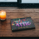 A spiral notebook with a colorful cover featuring the text 'COSMIC ATTIC' and 'Mind Body Soul' in a stylized font, surrounded by a pattern of stars and abstract shapes in vibrant colors.